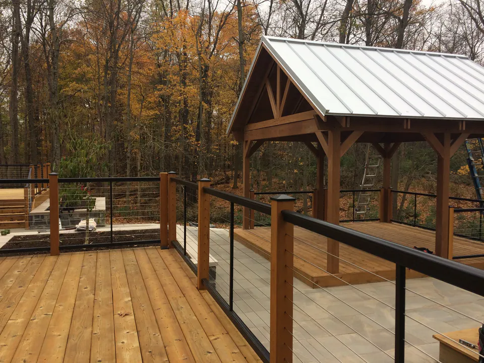 Cedar deck with cable railings leading to a timber pavilion with a metal roof in autumn woods