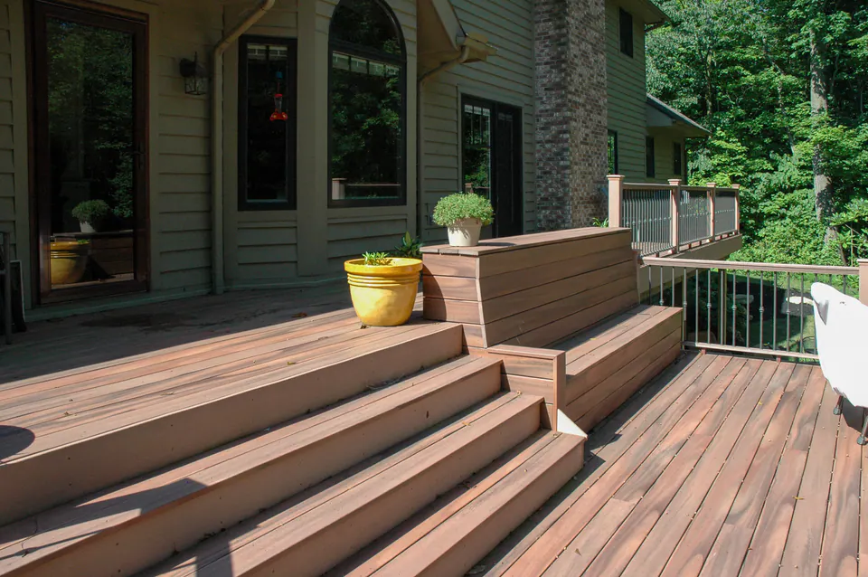 Wood-tone deck with broad stairs, built-in bench edge, black metal railing, planter pots, and wooded backyard view