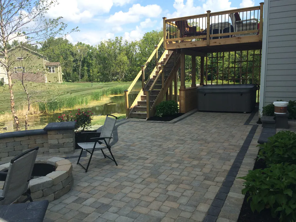 Raised wood deck with stairs down to a paver patio with a firepit and seat wall by a pond