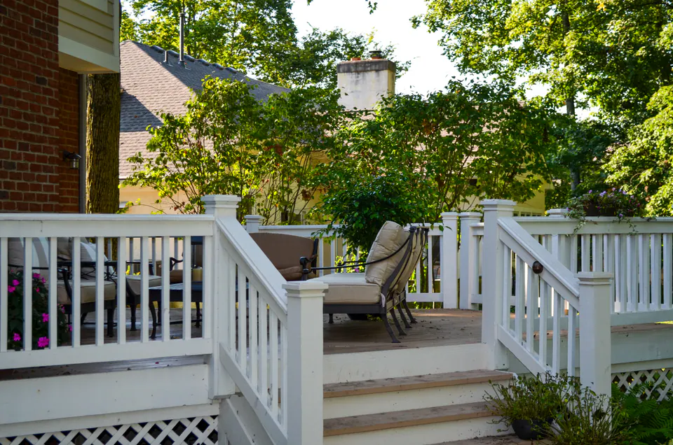 White deck railing with stair landing, outdoor seating, planter pots, lattice skirt, and wooded garden backdrop