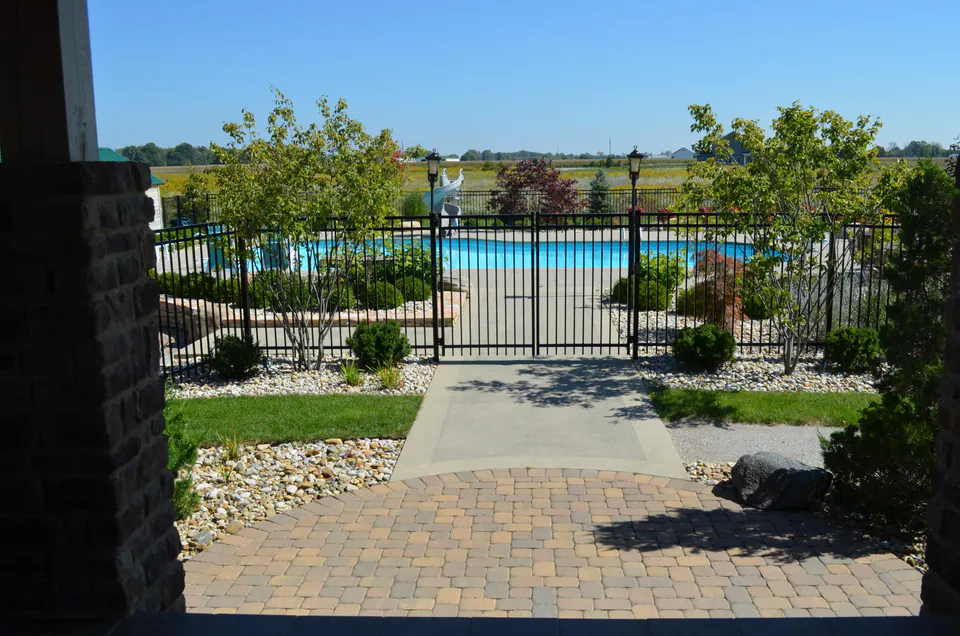 Black metal pool fence and gate across a paver walk, with planting beds on both sides and the pool beyond