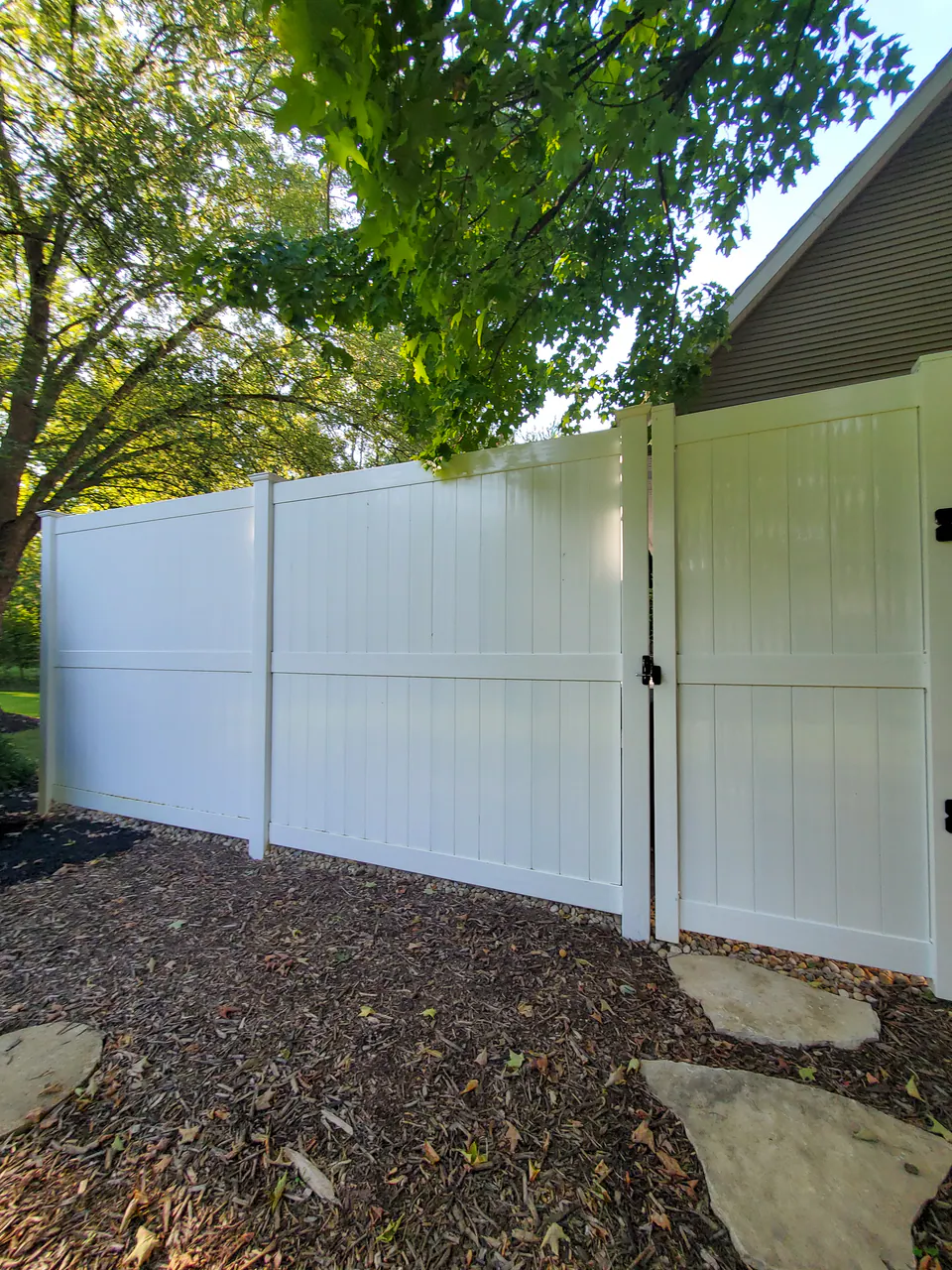 Tall white privacy fence and gate beside a house with mulch bed, stone step, and tree canopy