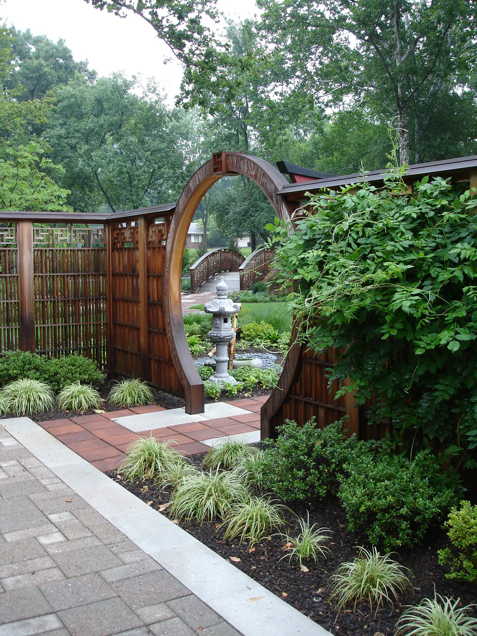 Round moon-gate opening in a wood privacy fence beside a paver walk with ornamental grasses