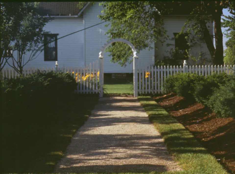 White picket fence and arched arbor along a gravel path beside a white farmhouse