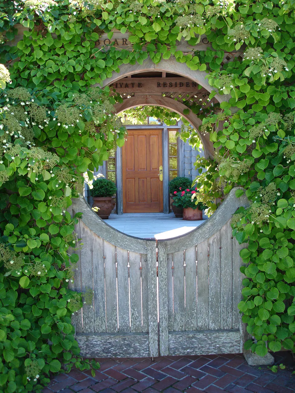 Wooden gate under a round opening in an ivy-covered wall, with a brick path leading to a front door