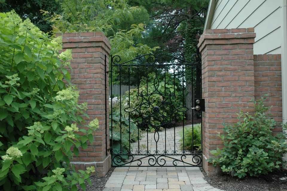 Ornate black iron garden gate between brick piers opening to a paver path