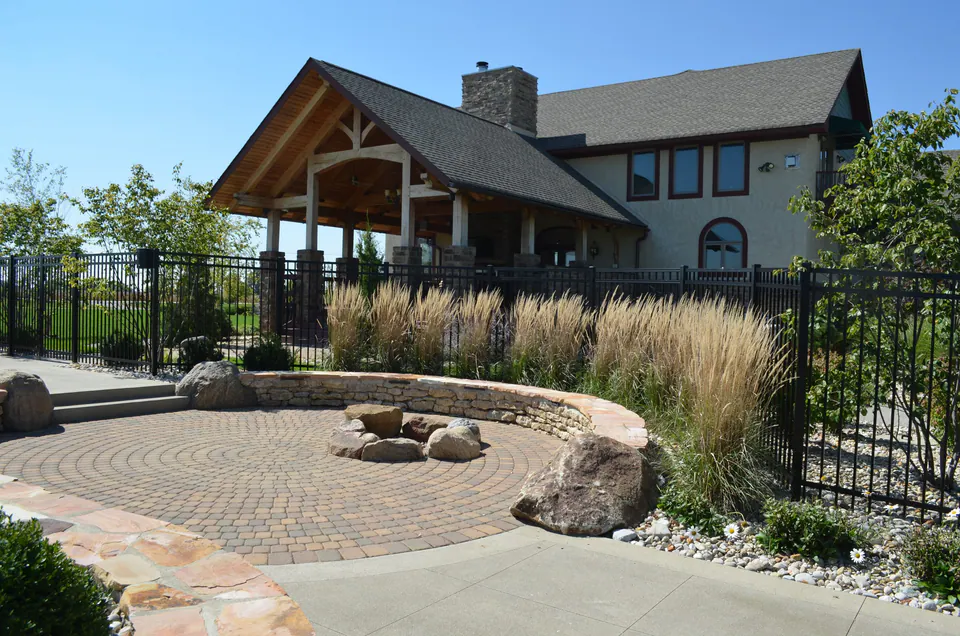 Boulder fire ring on a circular paver patio with a low stone wall and grasses beside a pool fence
