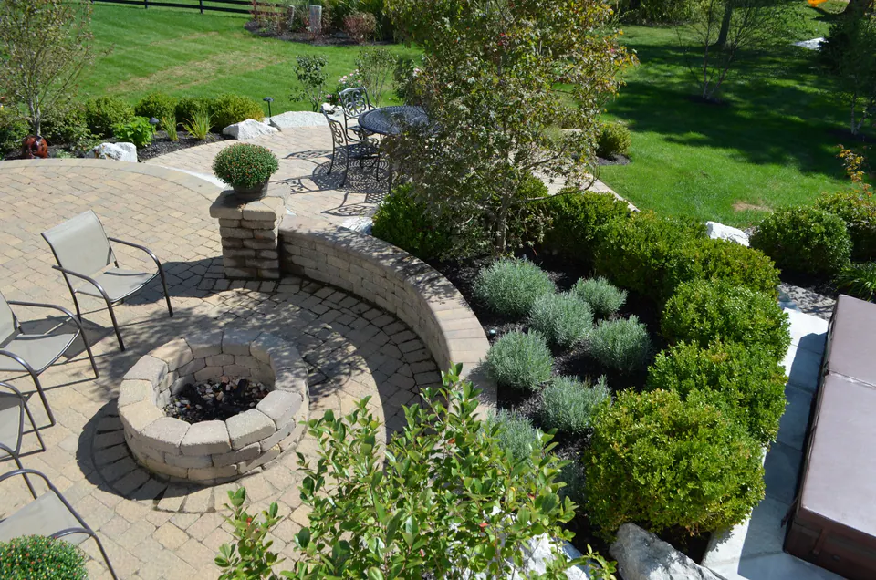Round block firepit on a paver patio below a curved seat wall and shrub garden