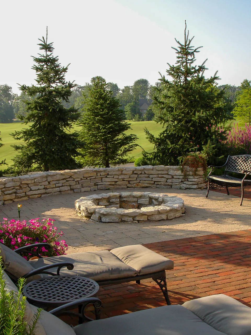 Round natural stone firepit on a brick patio with evergreen trees, seating, and open lawn beyond