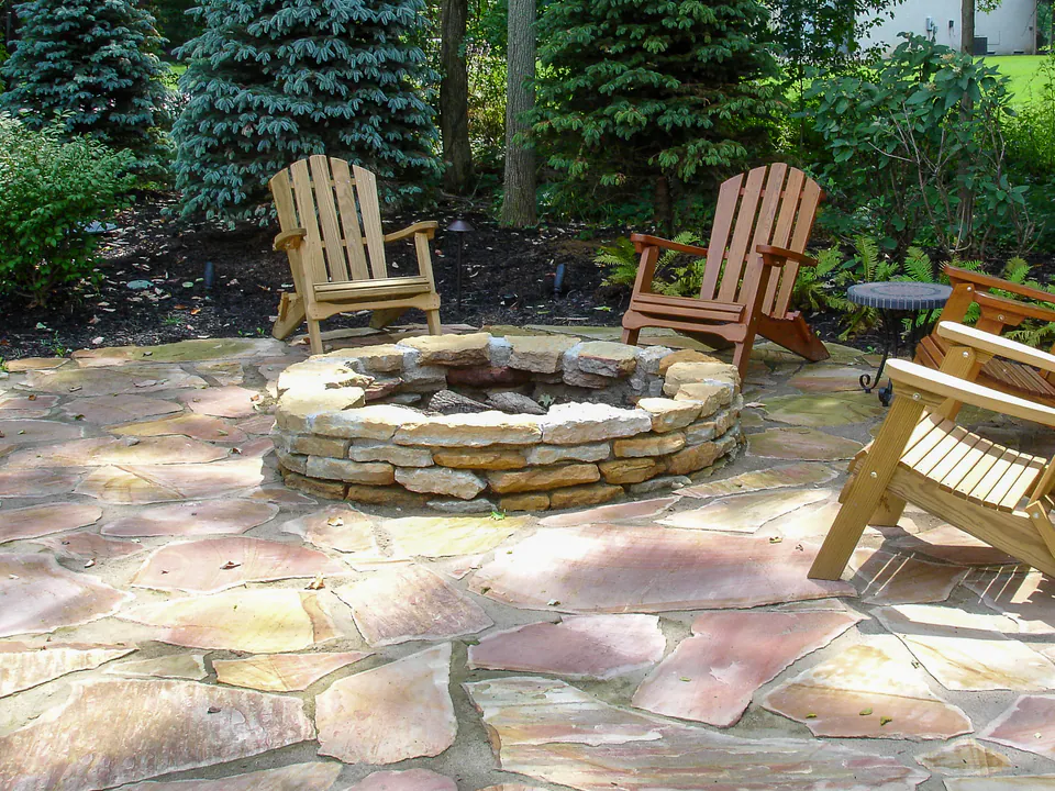 Round natural stone firepit on irregular flagstone with Adirondack chairs and woodland planting