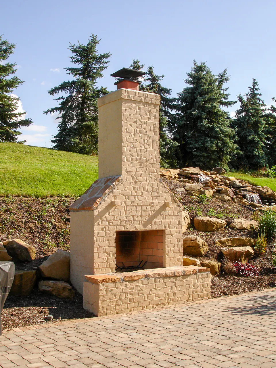 Painted brick outdoor fireplace on a paver patio with boulders, evergreen trees, planted slope, and waterfall beyond