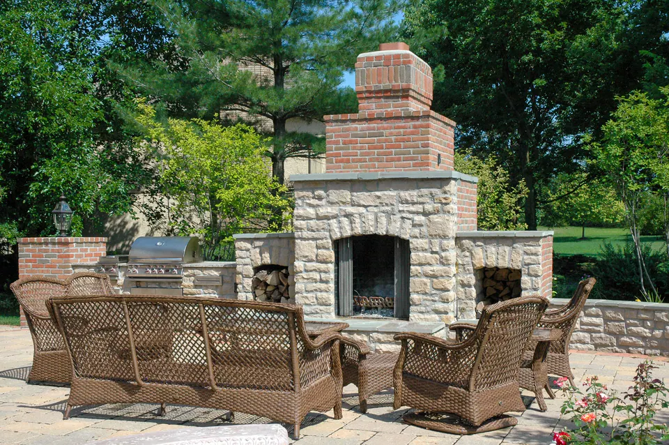 Brick and limestone outdoor fireplace with cut stone caps, wood storage, stainless grill, and paver patio seating