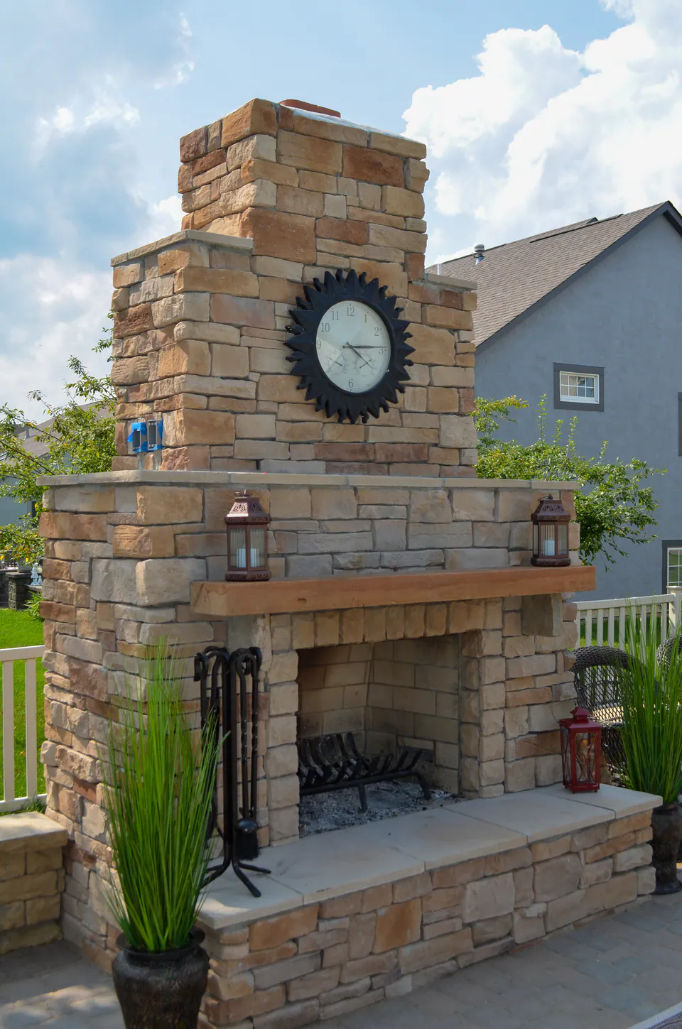 Natural stone outdoor fireplace with wood mantel, clock, stone hearth, fire tools, paver patio, planter pots, and privacy hedge