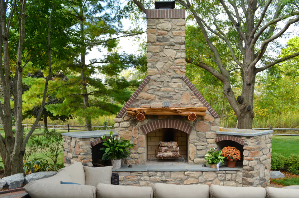 Natural stone outdoor fireplace with arched firebox, stone hearth, wood storage, patio seating, planter pots, and wooded backdrop