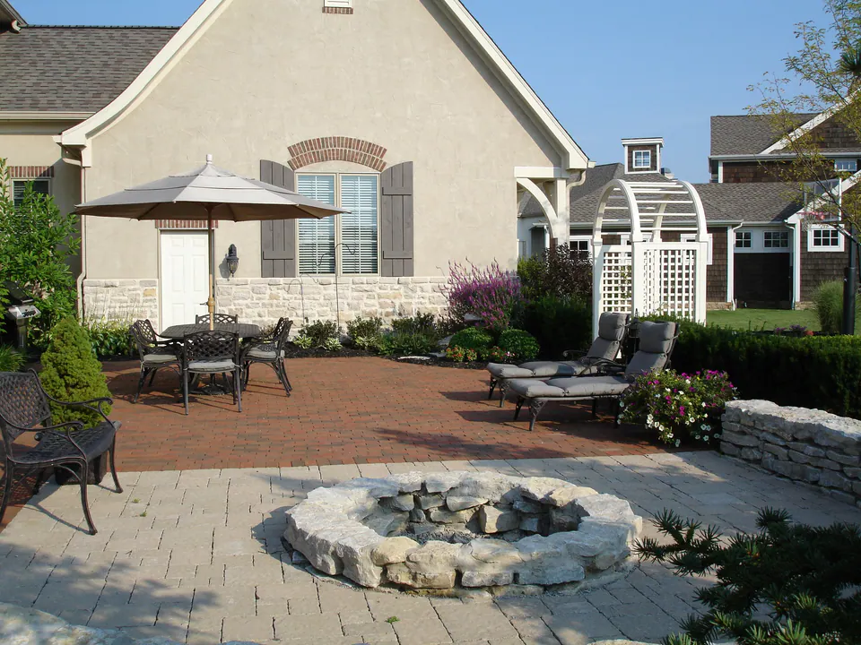 Round natural stone firepit on a paver patio with dining area and white arbor behind