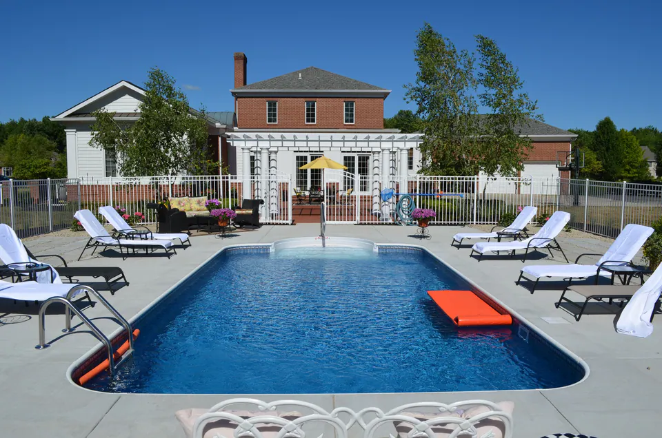 Large rectangular pool with a wide concrete deck, lounge chairs, metal fence and a white pergola