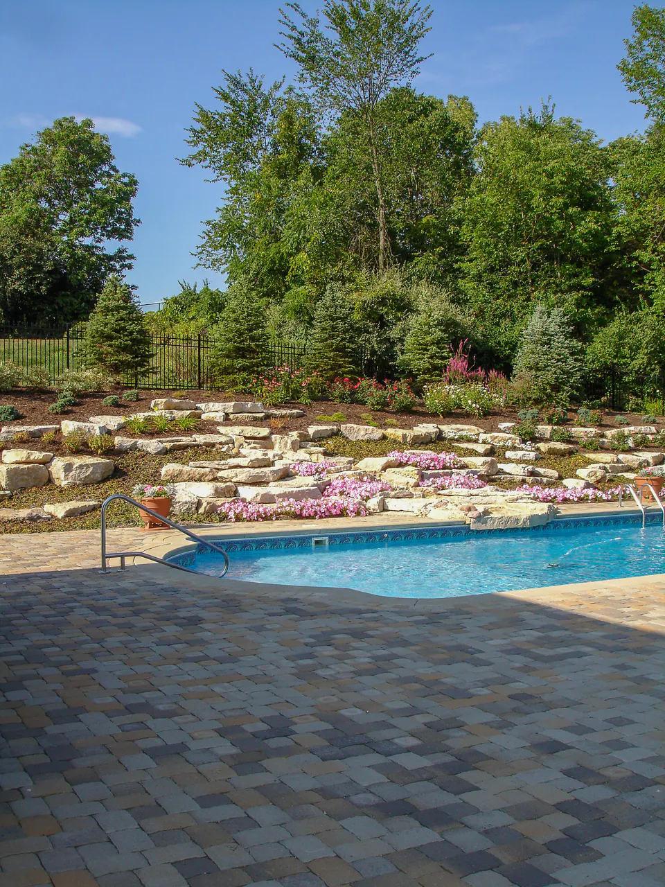 Pool with paver deck, natural limestone boulder slope, flowering planting, evergreen screening, and black metal fence.
