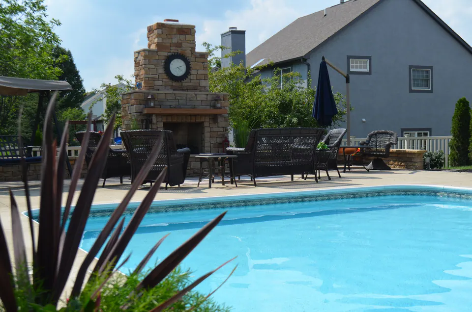 Pool in the foreground with a stone outdoor fireplace and patio seating behind it