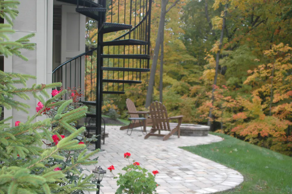 Curved paver patio at the foot of a black spiral stair, with Adirondack chairs and fall woods