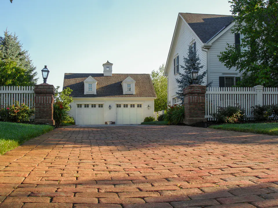 Brick paver driveway leading through stone gate piers toward garage doors with lawn and planting beds