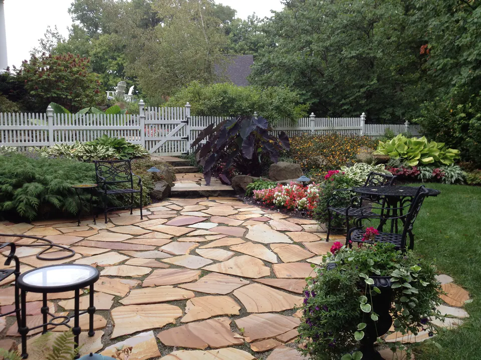 Irregular flagstone patio with table and chairs, colorful planting and a white picket gate
