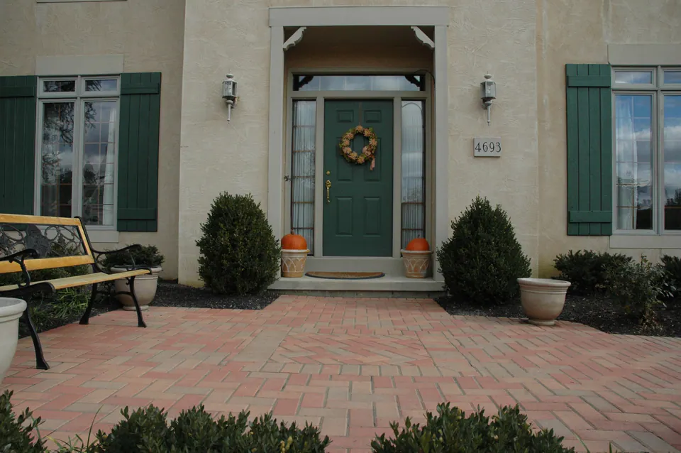 Front entry court in herringbone pavers with a bench, clipped evergreens and pumpkins at the door