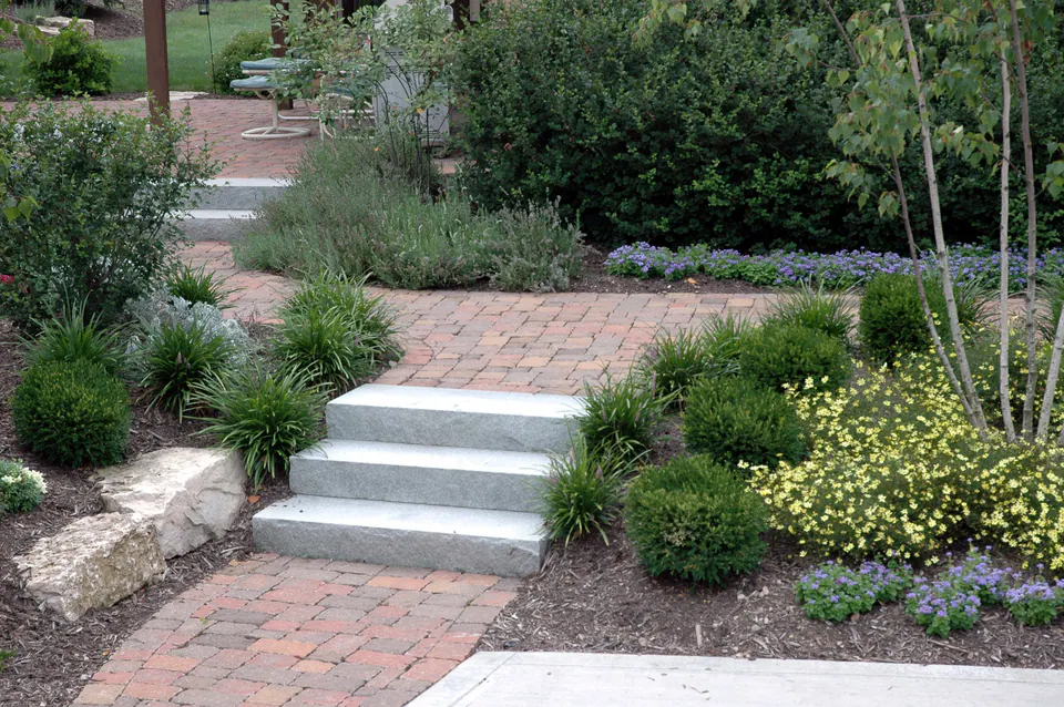 Three granite slab steps in a brick paver garden walk with boulders and perennials