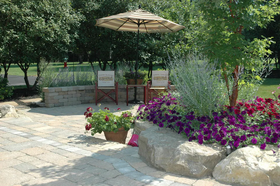 Corner of a paver patio with boulders, purple petunias, grasses and a planted pot