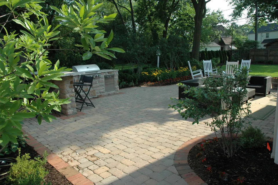 Paver patio with a stone grill island, Adirondack chairs and a small tree in a planting bed