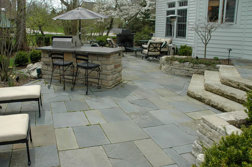 Full-range bluestone patio with stone outdoor kitchen, seating, low planting walls, and natural stone steps
