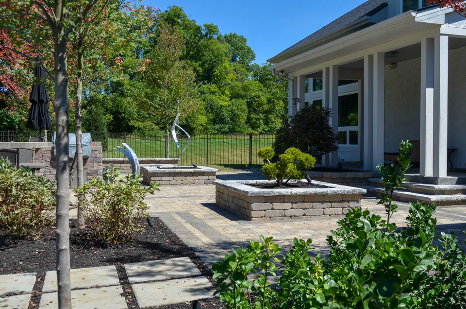 Paver courtyard patio with raised planting beds, stone caps, porch columns, sculpture, and black metal fence