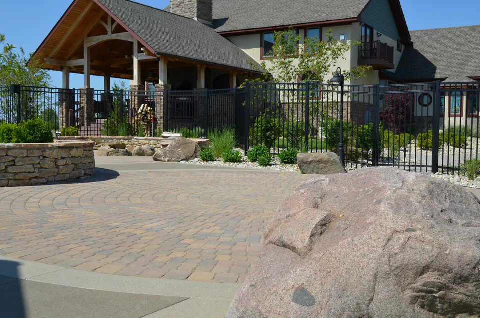 Paver motor court with a large boulder, stone planter and black metal fence at a house