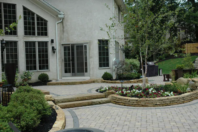 Paver patio with a raised stone planter full of annuals, stone steps and a young tree