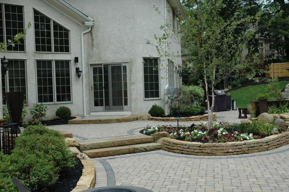 Paver patio with a raised stone planter full of annuals, stone steps and a young tree