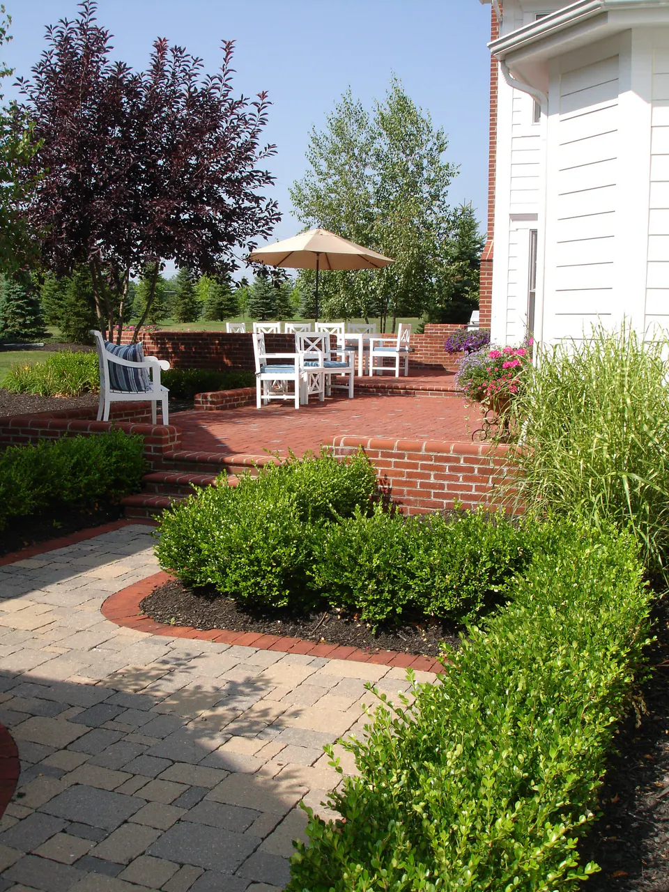 Paver walk bordered by clipped boxwood hedges leading to a brick patio with dining table