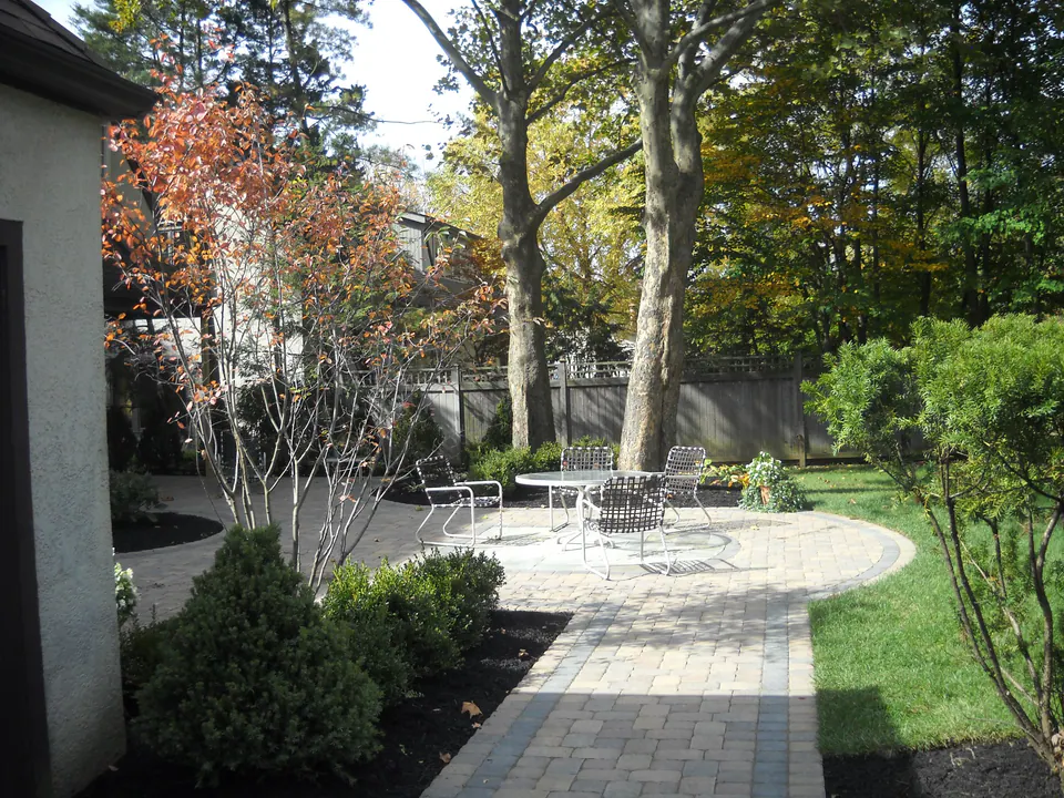 Paver walkway along a house leading to a round patio with a table under tall trees