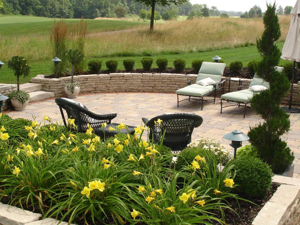 Round paver patio with a stone seat wall, lounge chairs and yellow daylilies overlooking a golf course