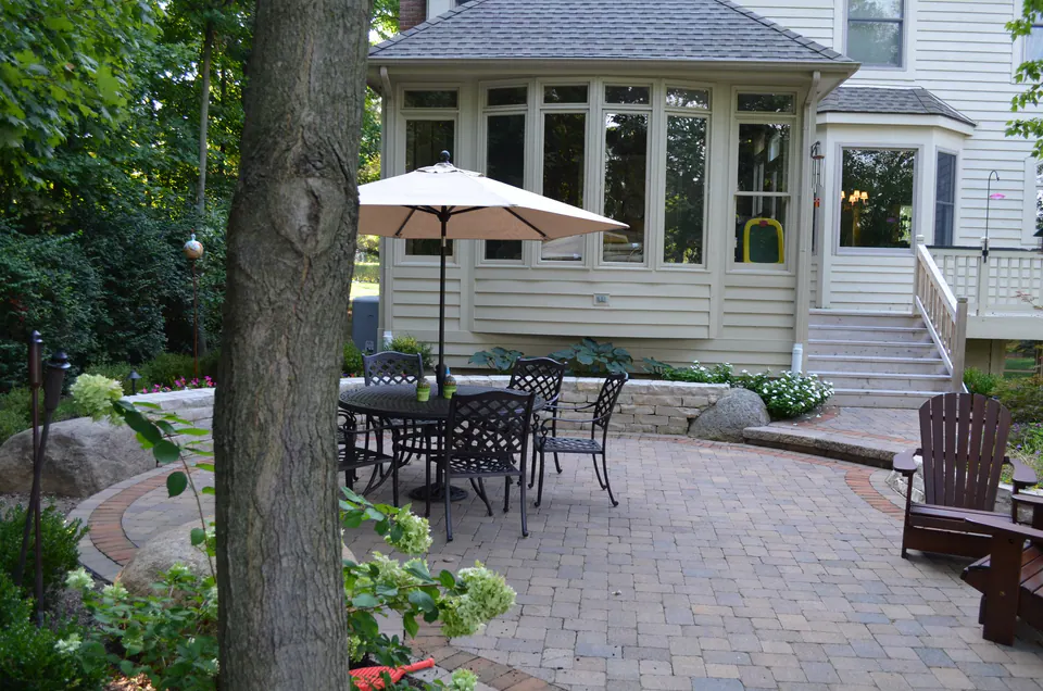 Round paver patio with dining table and umbrella below a sunroom, edged with boulders and hydrangeas