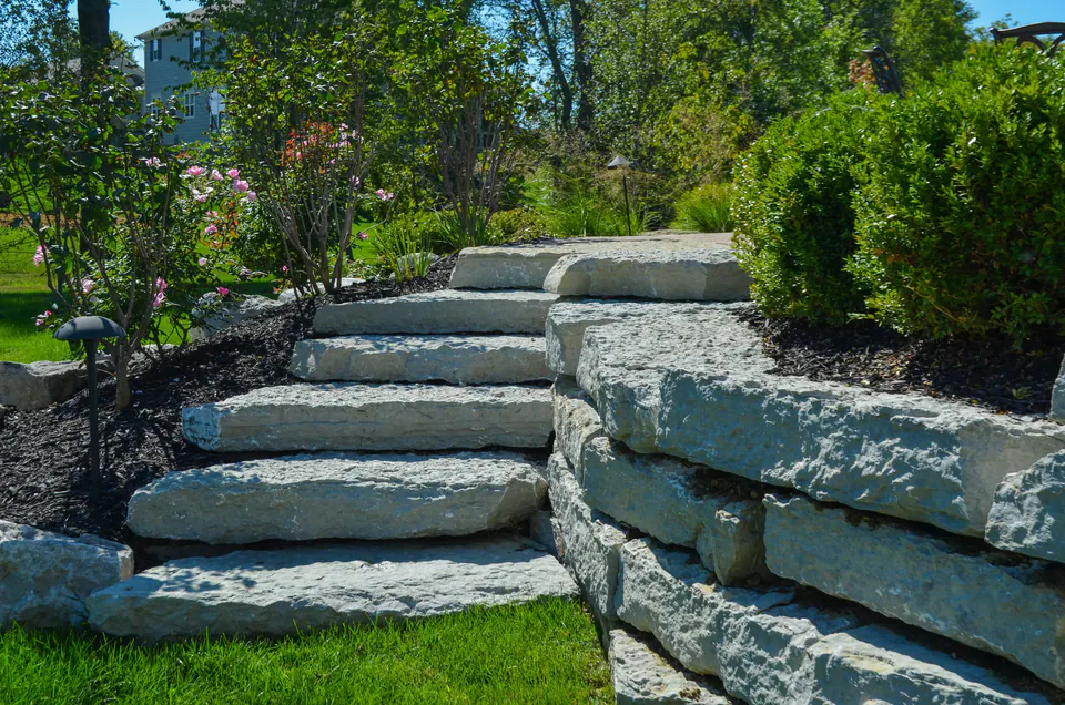 Large limestone outcropping steps beside a boulder wall, roses, shrubs, and black mulch