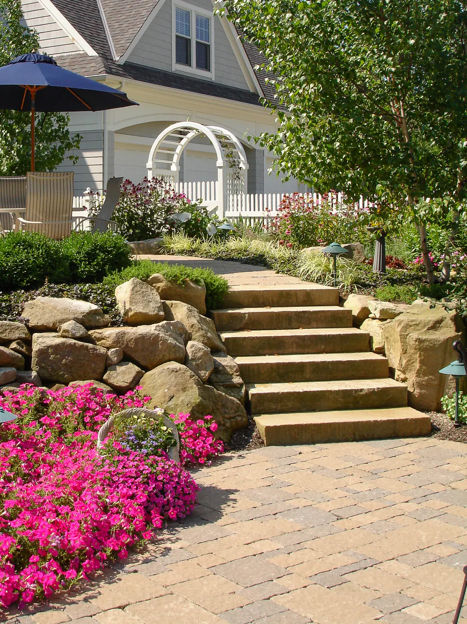 Limestone outcropping steps between flowering beds, boulders, paver walk, and white garden arbor