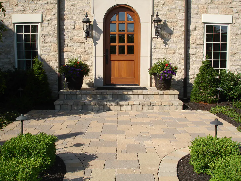 Wide stone steps and a paver court at an arched wooden front door with urns and lights