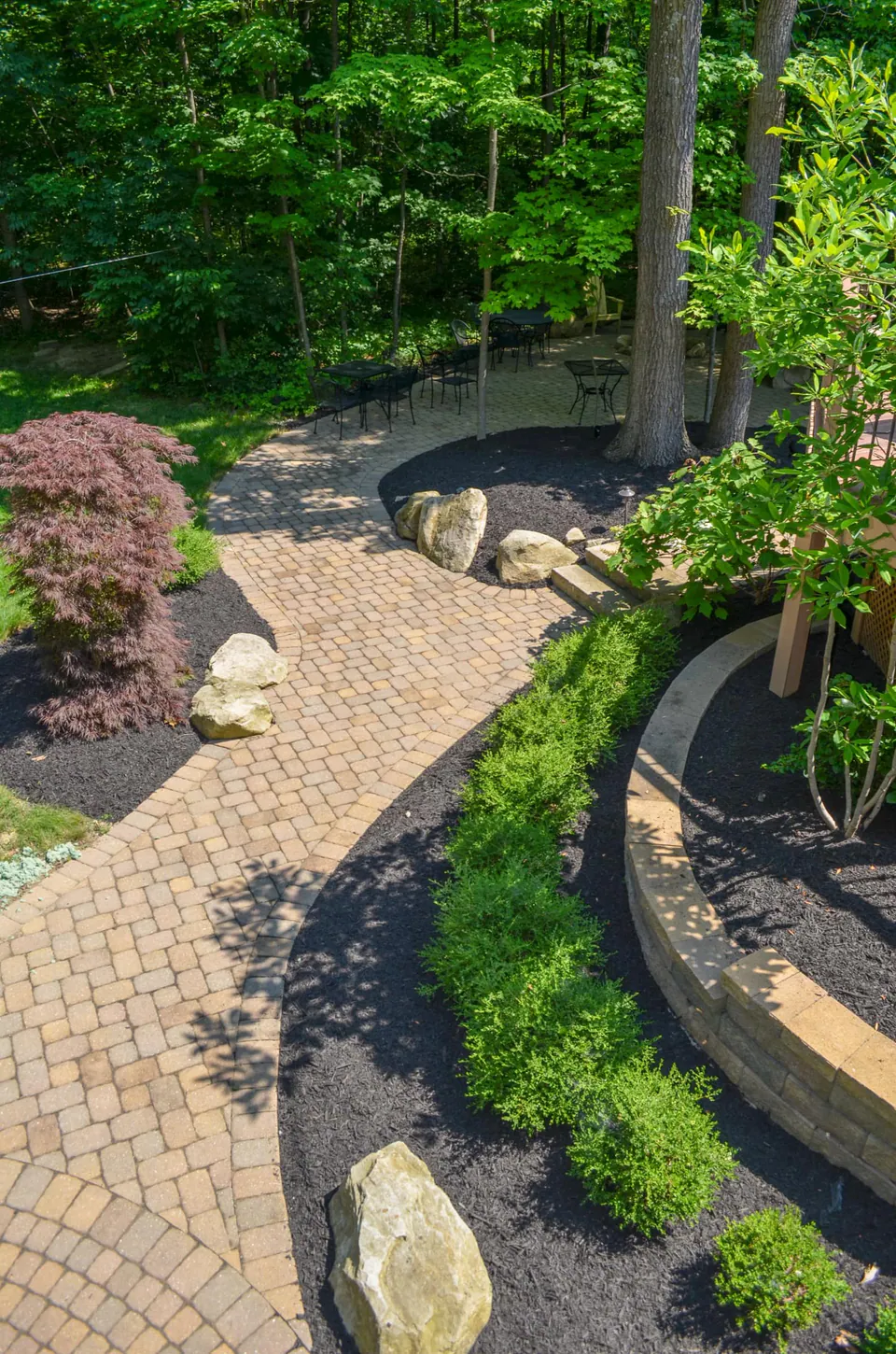 Curving paver walkway through a wooded backyard with boulders, mulch beds, low wall edging, and patio seating