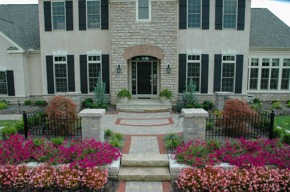 Formal front walk with concrete pavers, brick accent border, cut stone steps, black metal fence, and seasonal planting