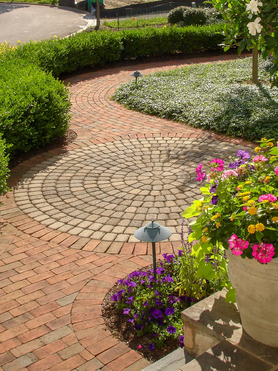 Circular paver medallion with clay brick border, cobble-style center, boxwood hedge, groundcover, and seasonal flowers