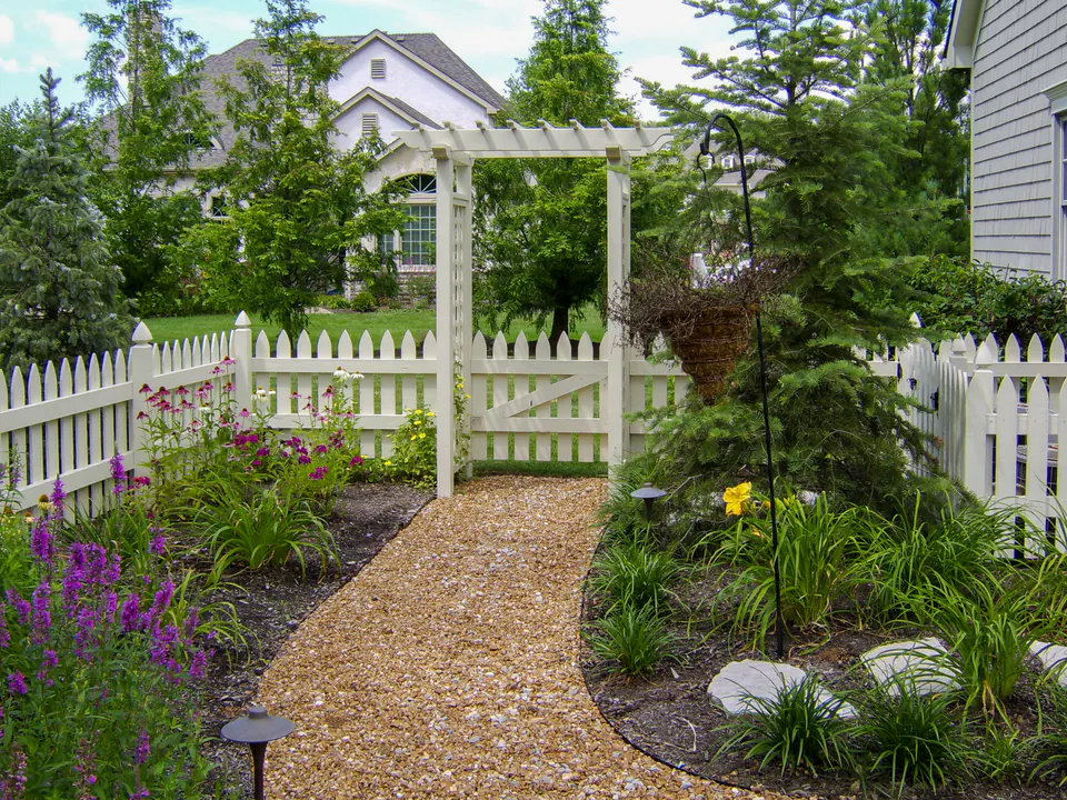 White garden arbor and picket fence over a gravel garden walk with perennial planting and path lighting