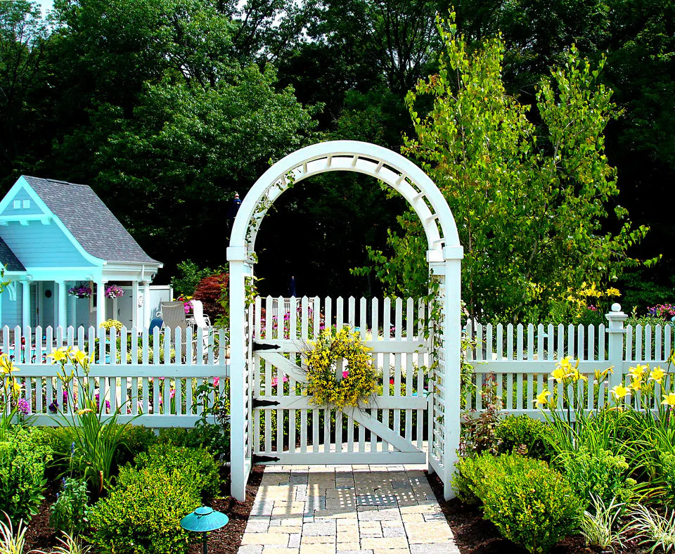 White arched arbor with a picket gate and wreath in a white picket fence, with a paver walk and perennial beds