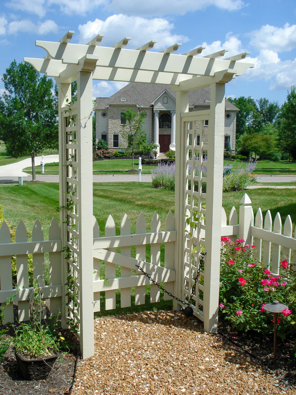 White garden arbor and picket fence gate with climbing vines, rose planting, and gravel entry