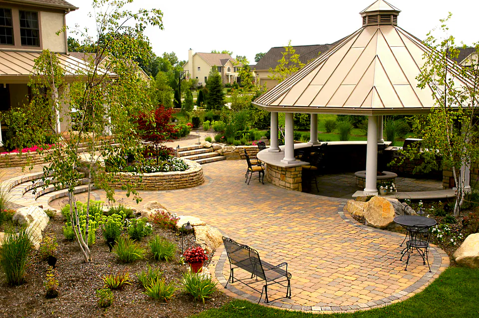 Round copper-roofed gazebo at the edge of a circular paver patio with tiered planting beds and a stone wall