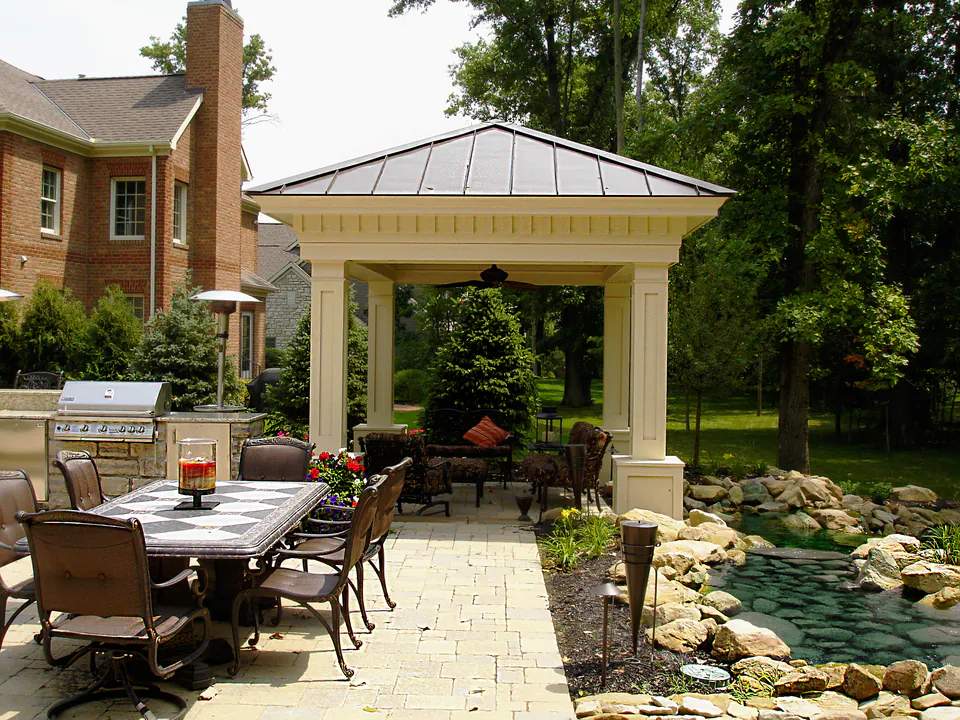 Open pavilion with a hip roof over an outdoor kitchen and dining table on a paver patio beside a pond
