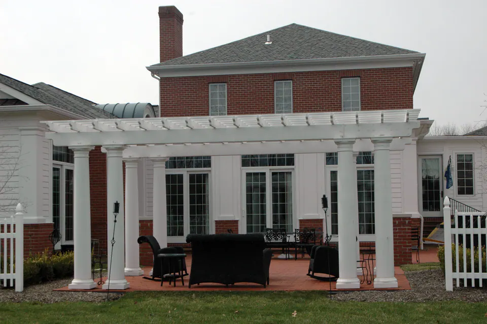 White columned pergola across the back of a brick house over a brick patio with wicker furniture